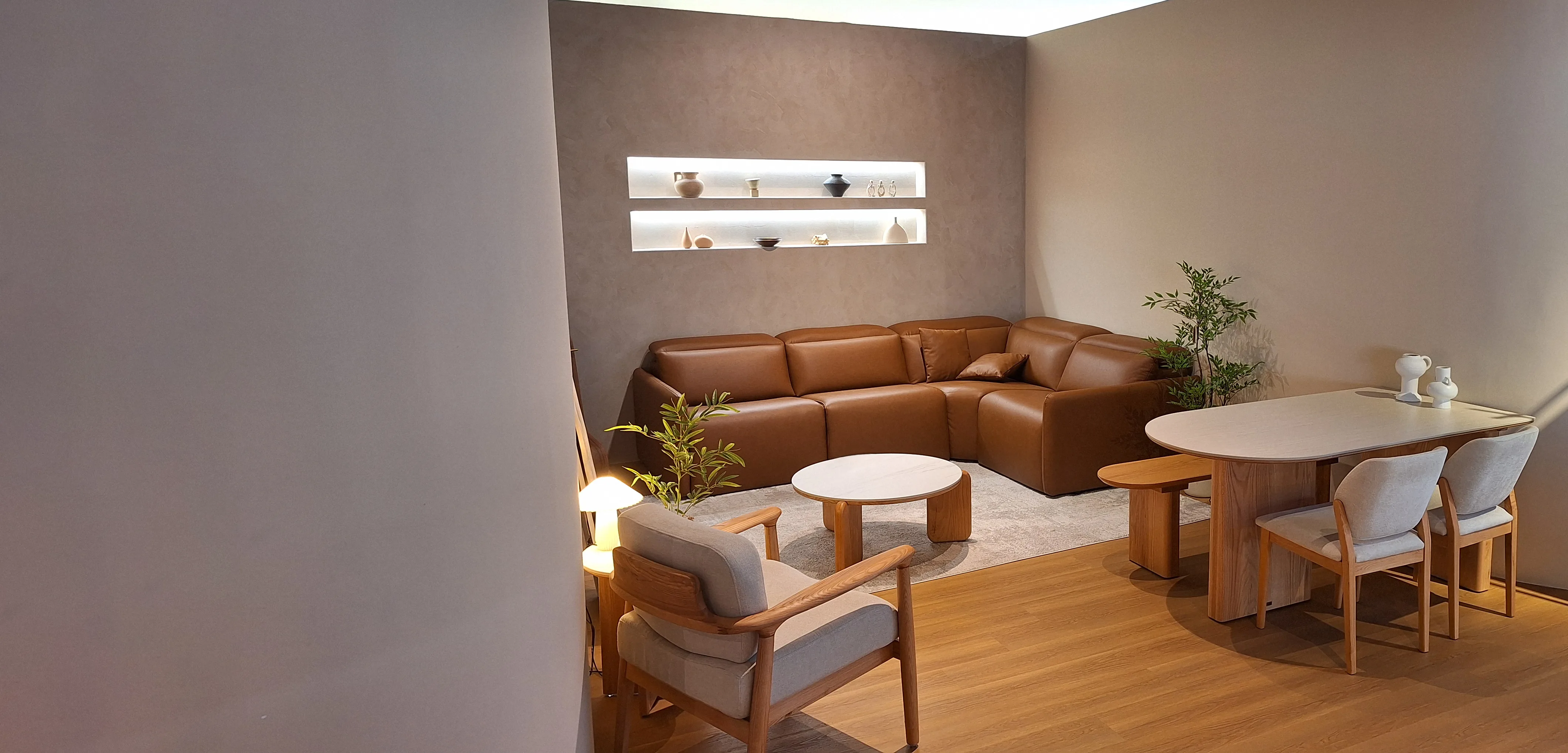 A Son&Bear showroom room set: a tan leather corner sofa against a plaster wall with a lit display niche, an oak dining table and chairs to the right, and a cane-and-oak armchair in the foreground.